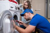 Handwerker im Blaumann schraubt an Waschmaschine, dahinter die beunruhigte Besitzerin &copy; by Grustock/AdobeStock